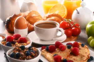White cup filled with tea surrounded by fresh fruit and oats, boiled egg, cheery tomatoes, fresh oranges and orange Juice, a bagel and croissant 